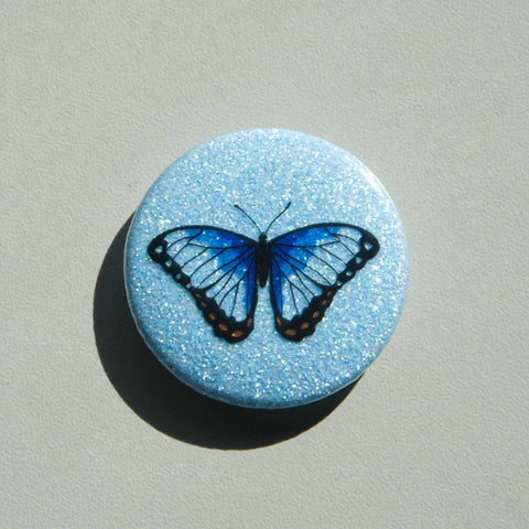 A blue sparkly magnet featuring an ble morpho butterfly on a white table.