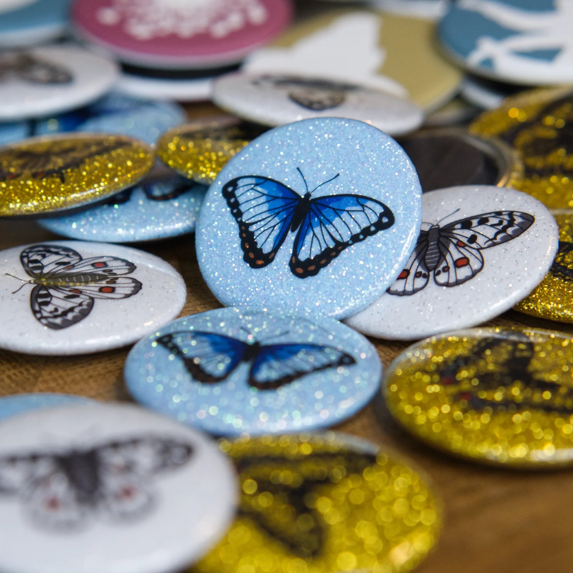 A blue sparkly magnet featuring an blue morpho butterfly leans against other colourful magnets on a wooden table.