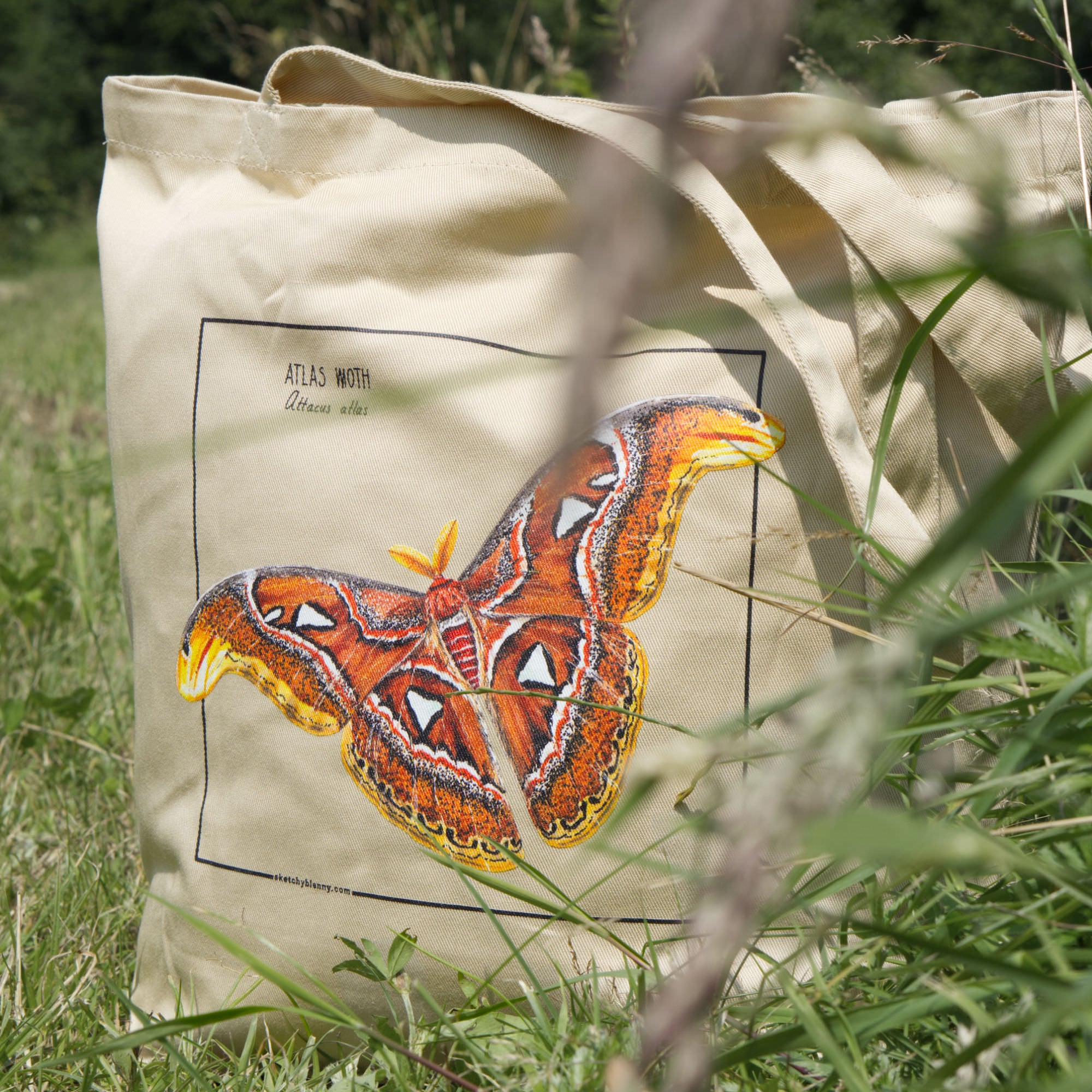 Product photo of our atlas moth organic tote bag. Here seen standing in high grass outside.