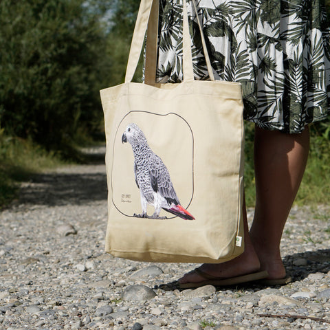 Our tote bag with a model outside. This sturdy bag comes with a beautiful print of a hand-drawn juvenile grey parrot. 100% organic cotton. Beach bag, shopping bag, travel bag, or just as a present. PETA-approved vegan.