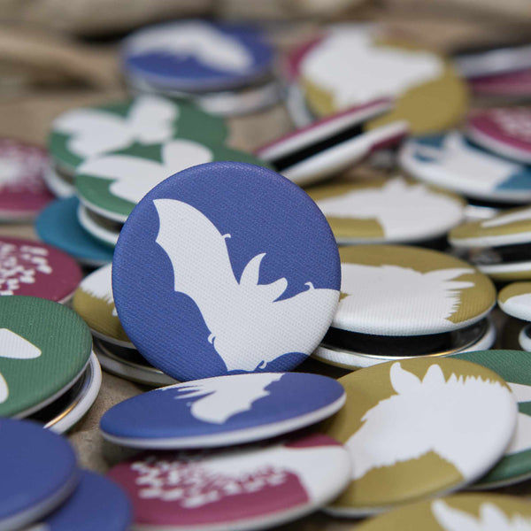 A navy-blue, fabric-covered magnet with the silhouette of a Brown Long-Eared Bat leans against a whole bunch of other colourful magnets.