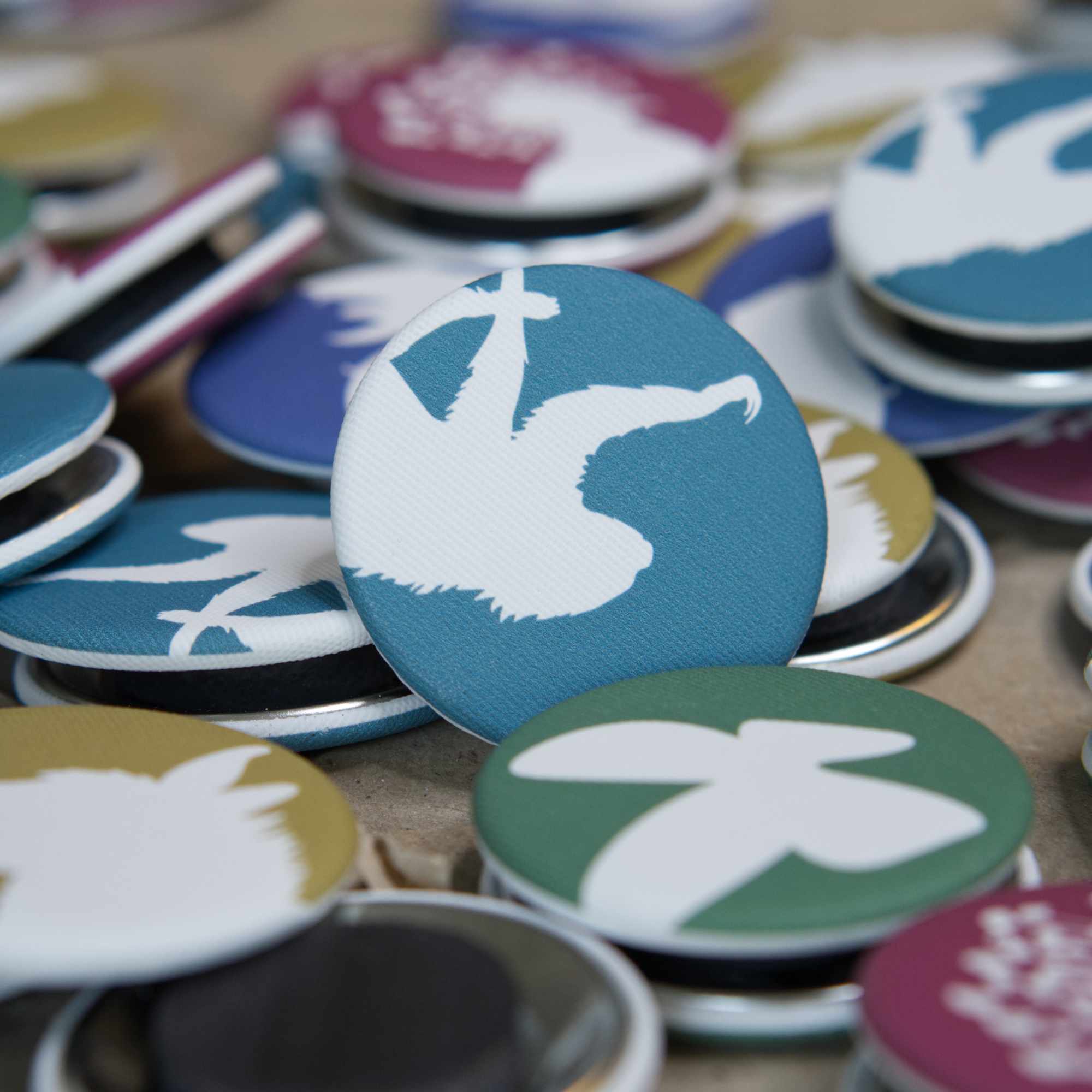 A teal-blue, fabric-covered magnet with the silhouette of a two-fingered sloth is lying on top of many other diferrent coloured magnets.