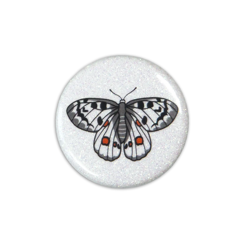 A white sparkly magnet featuring an apollo butterfly leans on a white. Top down photo.