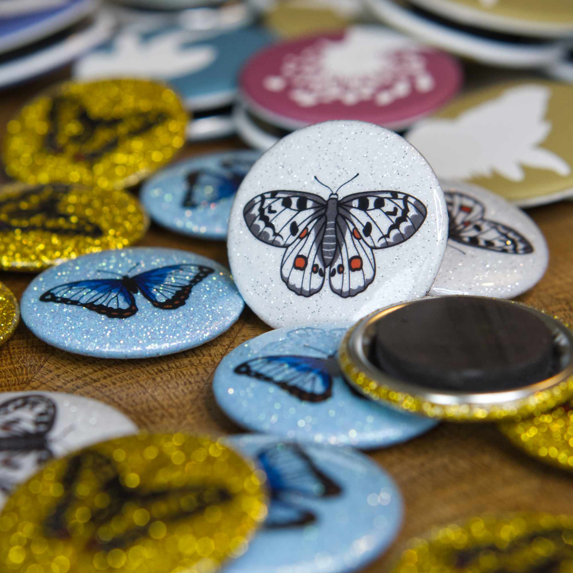 A white sparkly magnet featuring an apollo butterfly leans against other colourful magnets on a wooden table.