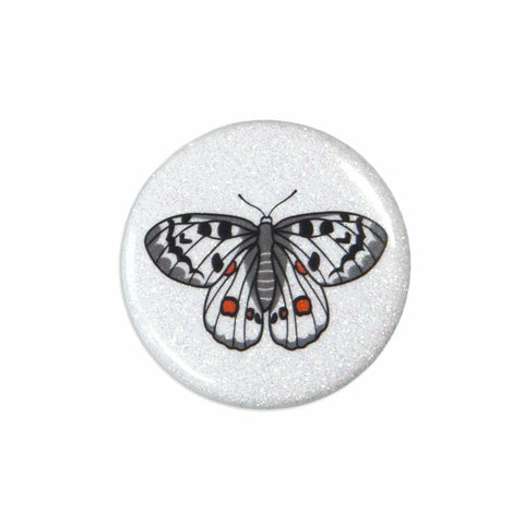 A white sparkly magnet featuring an apollo butterfly leans on a white surface.