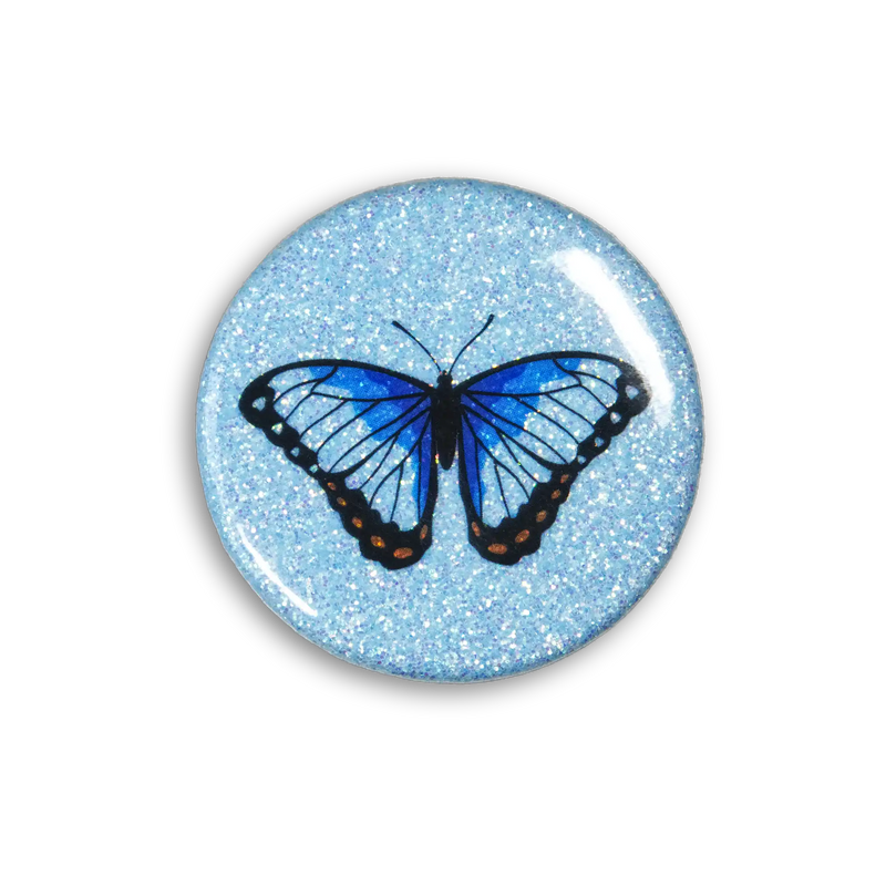 A blue sparkly magnet featuring an ble morpho butterfly on a white table. Top down photo.