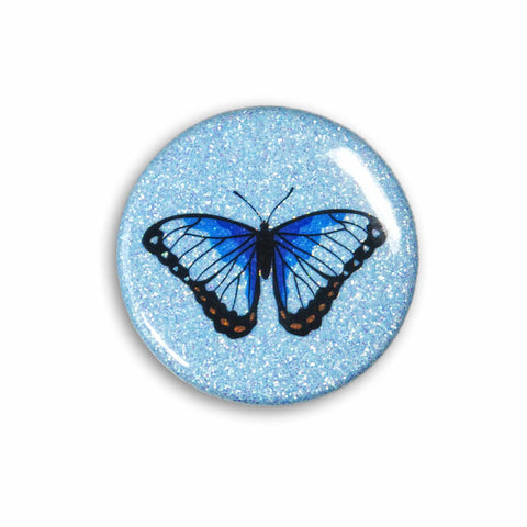 A blue sparkly magnet featuring an ble morpho butterfly on a white table.