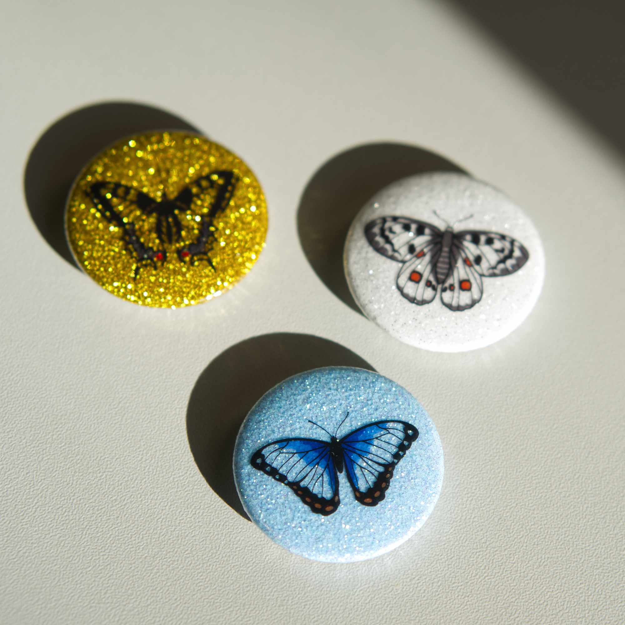 Three glittery magnets on a white surface, one is golden, one blue and one white.