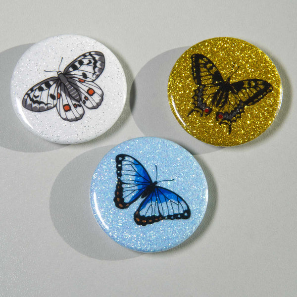 Three glittery magnets on a white surface, one is golden, one blue and one white.