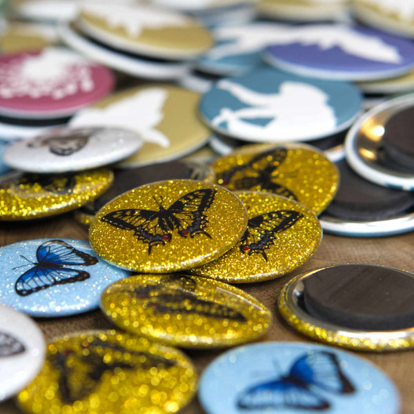 Several beautiful gold-glitter swallowtail magnets lie scattered amongst other colourful magnets on a wooden table.