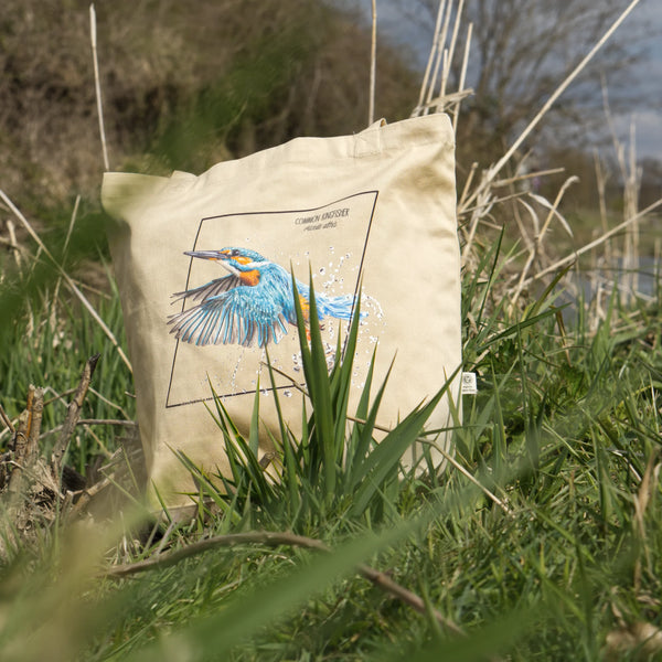 Organic tote bag featuring a common kingfisher in an outside photoshoot, standing amongst long grass along a river.