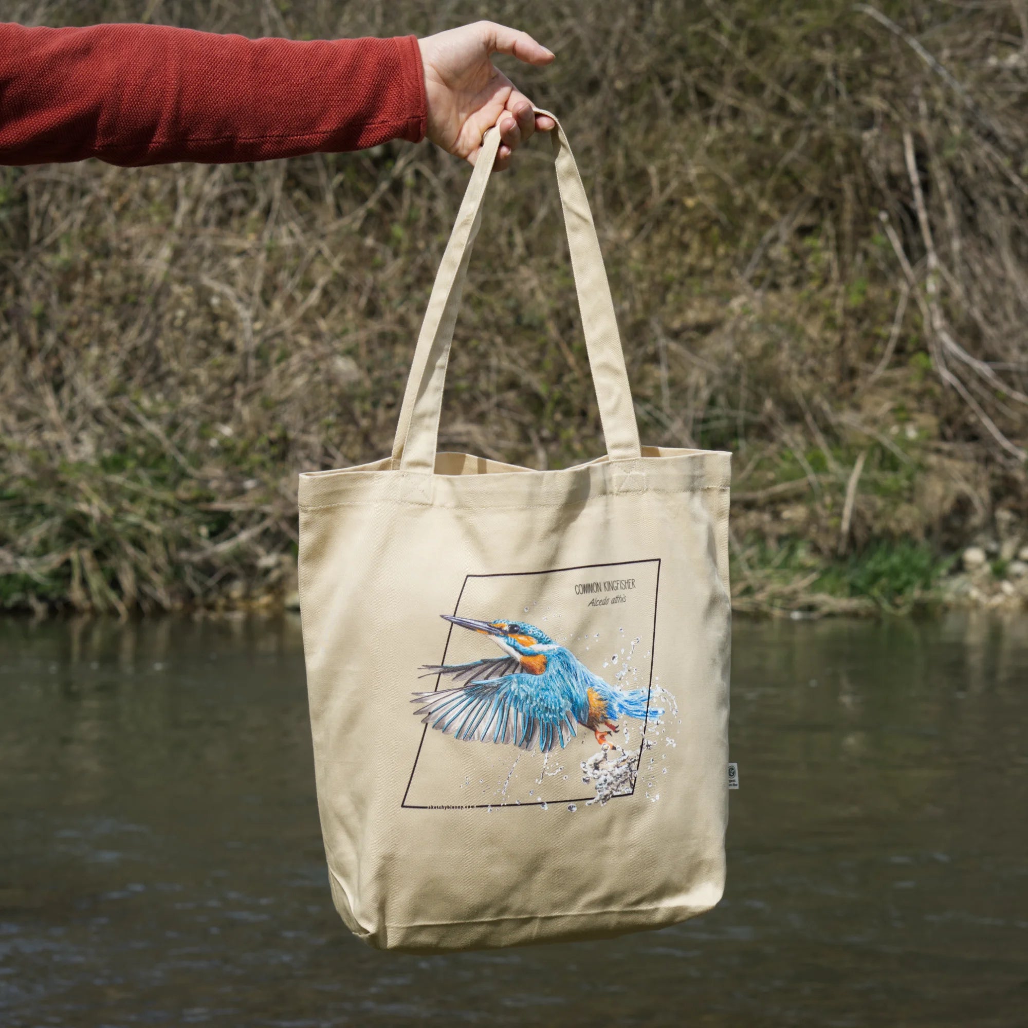 Organic tote bag featuring a common kingfisher in an outside photoshoot, hanging from the hand of a male model over a river.