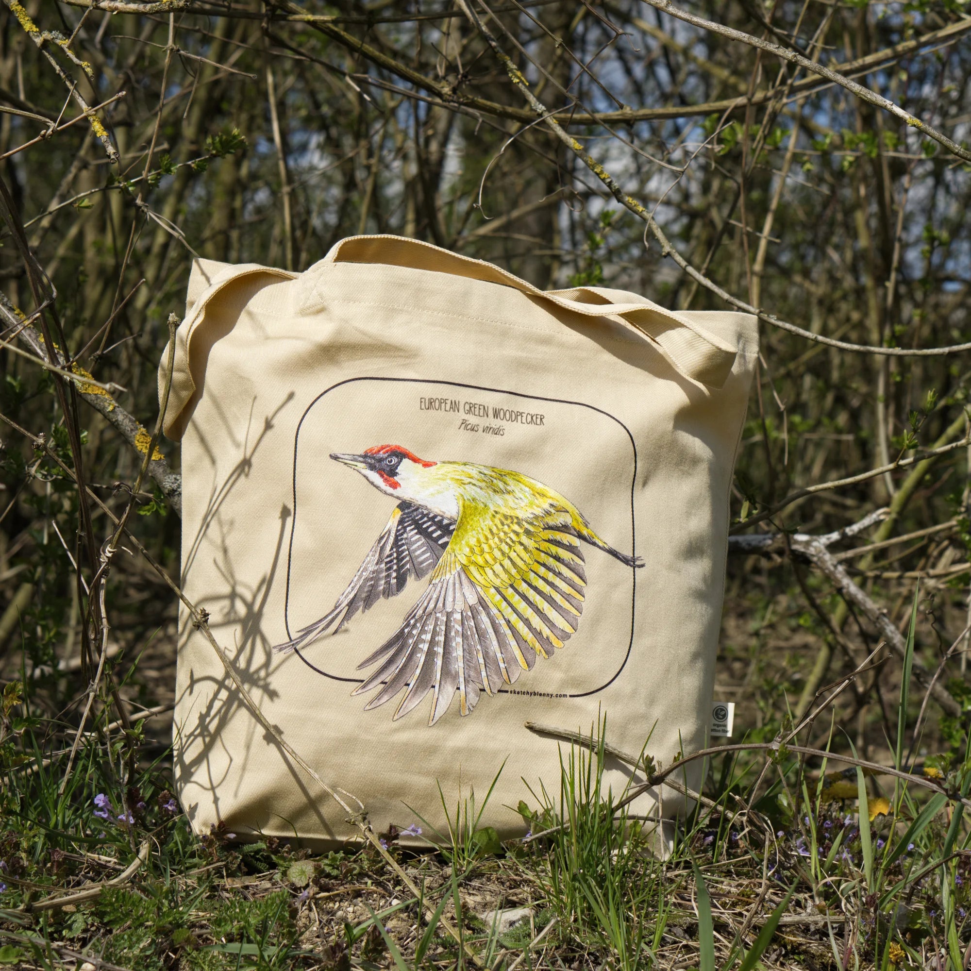 A European green woodpecker print on a 100% organic tote bag. The photo shows the bag standing in nature amongst branches
