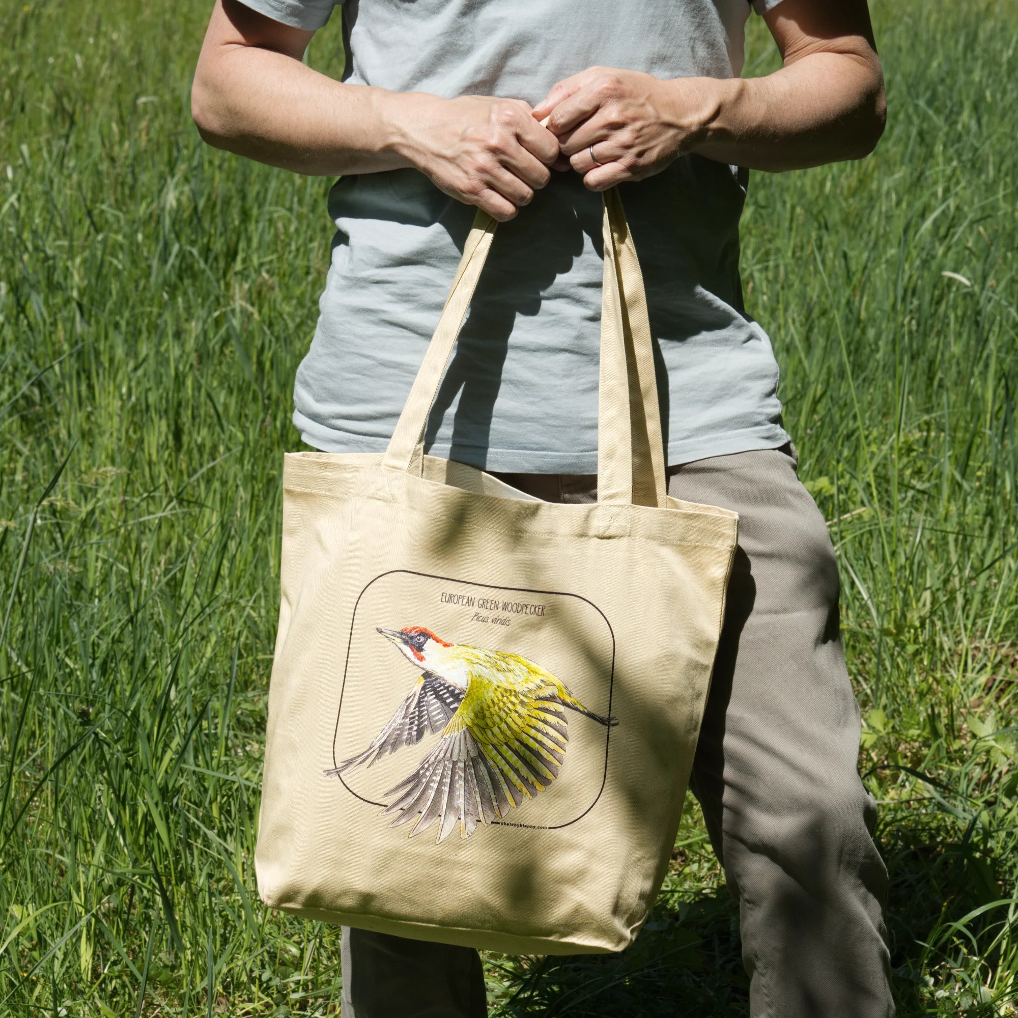 A photo of a model holding a tote bag outside in nature. European green woodpecker print on a 100% organic tote bag.