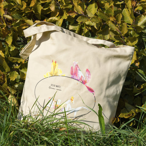 Photo of our  orchid mantis 100% organic cotton tote bag outside on grass in front of colourful leaves. Three colourful insects are on the sturdy, spaciuos bag.