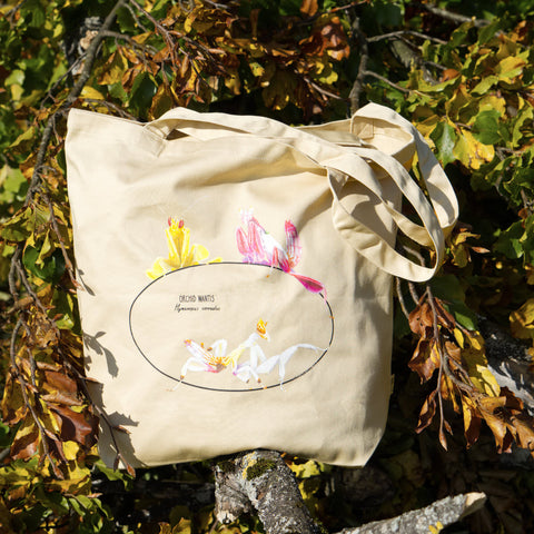 Photo of our  orchid mantis 100% organic cotton tote bag outside standing amongst colourful leaves. Three colourful insects are on the sturdy, spaciuos bag.