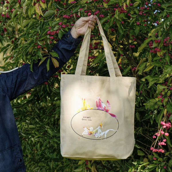 Photo of our  orchid mantis 100% organic cotton tote bag outside being held from the handle in front of a bush. Three colourful insects are depicted on the sturdy, spaciuos bag.