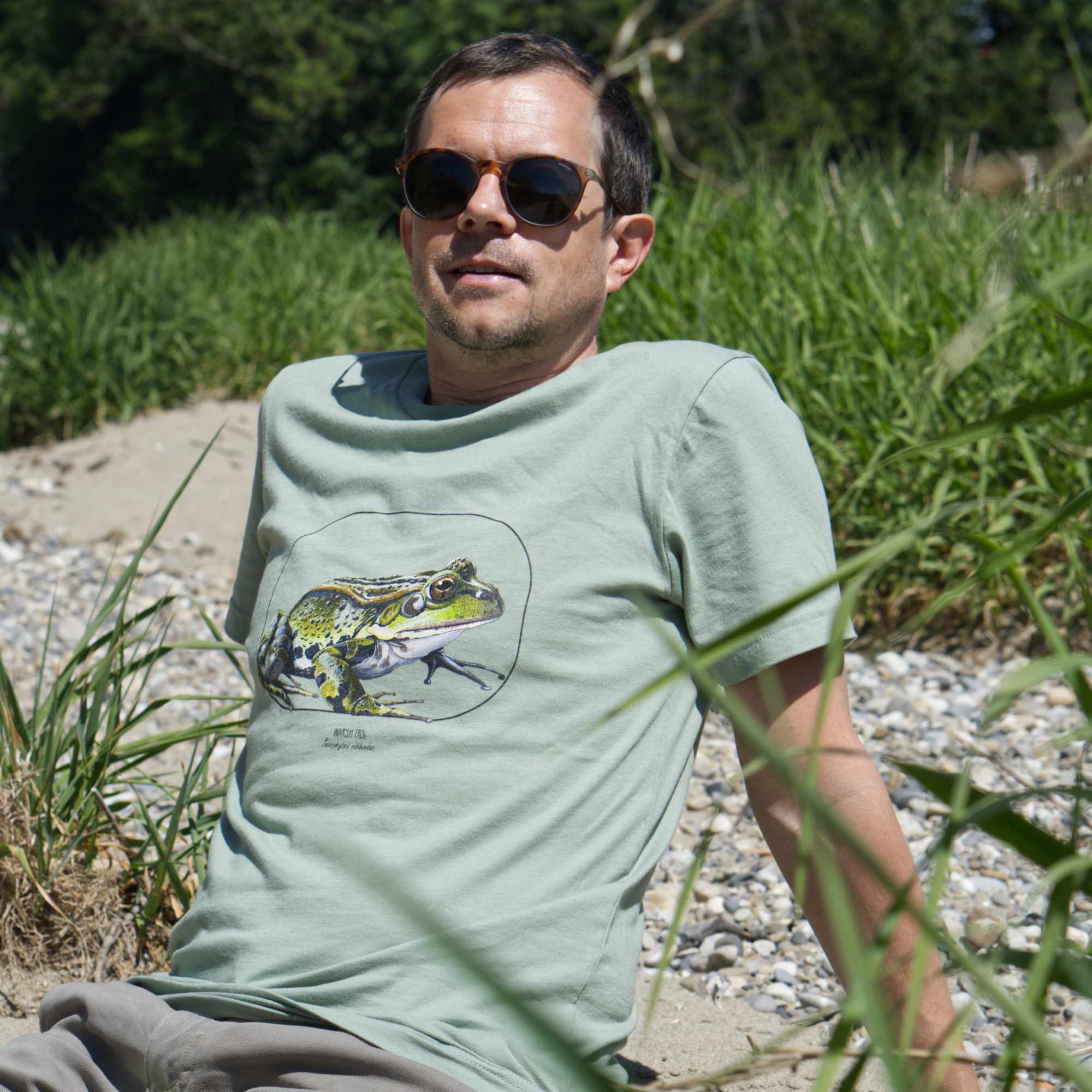 Photo of a male model sitting on a beach, wearing a sage coloured t-shirt with a beautiful marsh frog print.