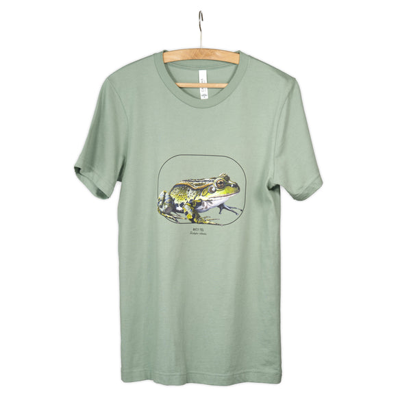 Hanging from a wooden hanger, this photo showcases our sage coloured t-shirt with a marsh frog print.