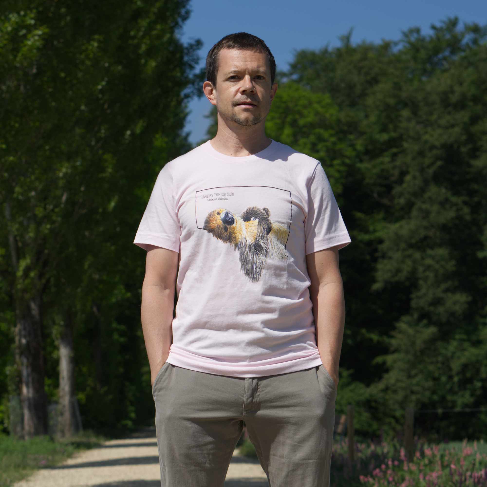 Outdoor photo of a male model standing and wearing the sift pink  linnaeus's two-toed sloth t-shirt. 